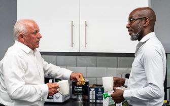 Two men stood in a kitchen holding drinks discussing mental health support. 
